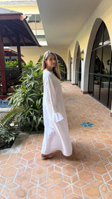 A woman wearing a long white kaftan dress stands in a tiled outdoor walkway with green plants and arched windows in the background.