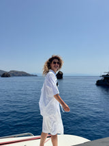 A woman with curly hair wearing a white short-sleeve shirt and matching shorts stands on the bow of a boat, looking back over her shoulder against a sunny blue sea and sky background.