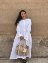A woman stands against a textured beige stucco wall wearing a long-sleeved white button-front tunic dress with voluminous sleeves and holding a woven beige tote bag.