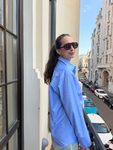 A woman wearing a light blue button-up shirt and black sunglasses stands on a balcony overlooking a city street with parked cars.
