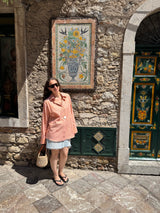 A woman wearing a light pink striped shirt and a light blue denim skirt stands on a stone patio in front of a rustic stone wall with a colorful floral tile mural and a green decorative door.