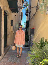 A woman stands in a narrow sunlit alleyway wearing a light peach-pink striped button-up shirt over a white mini skirt, with black sandals and a woven tote bag.