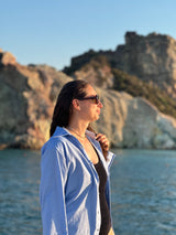 A woman in a light blue button-up shirt and sunglasses stands in profile by the water with rocky cliffs and blue sky in the background.
