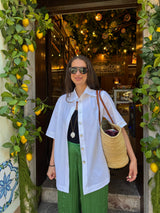 A woman wearing a white short-sleeve button-up shirt, green pleated trousers, and sunglasses stands in front of a decorative doorway adorned with lemon vines and warm hanging lights. She is carrying a woven straw tote bag.